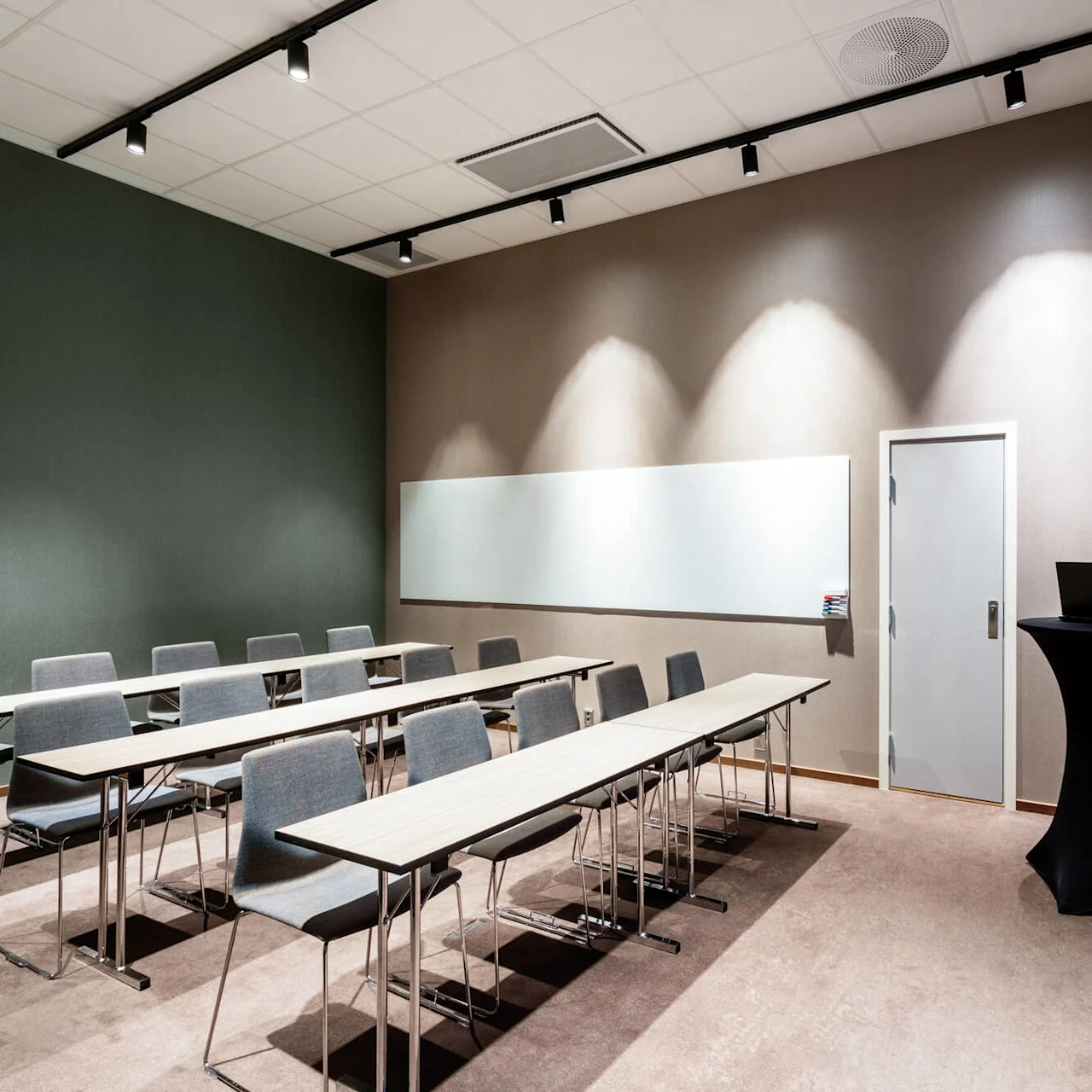 Classroom setup with rows of tables and chairs, facing a wall-mounted whiteboard. A small table with a laptop stands beside a closed door. Soft lighting enhances the study environment.