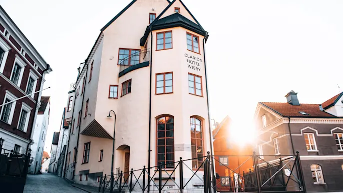 En høy, vinklet bygning med teksten "CLARION HOTEL WISBY" står på et gatehjørne under en solrik dag, omgitt av historiske strukturer med en varm glød fra solen.