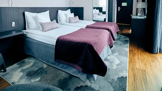 Two neatly made beds with purple quilted covers, situated in a modern hotel room. A large mirror, bedside table, and desk with a lamp complete the cozy space.