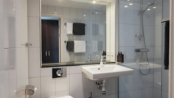 Ett ljust och modernt hotellbadrum med vitkaklade väggar, handfat, toalett och en duschplats med glasvägg.