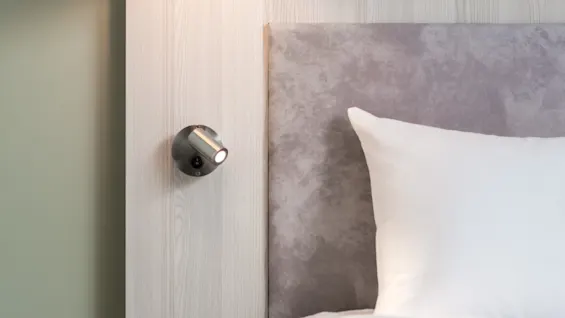 A wall light illuminates beside a bed with a gray upholstered headboard and white pillow, set against a light wooden wall.