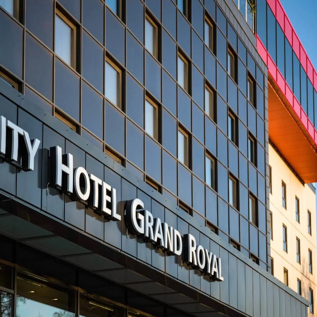 Quality Hotel Grand Royal-skilt vises på en moderne bygningsfacade med reflekterende vinduer, under en klar blå himmel.