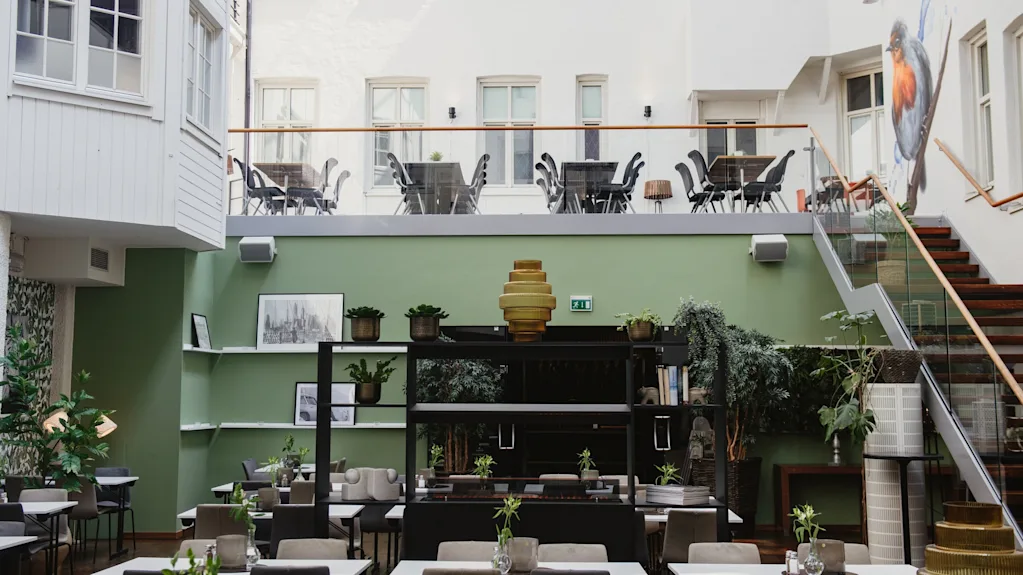 Restaurangbord med växter; omgivande grön inredning och konstverk skapar en modern, luftig atmosfär. Utomhusplatser på övervåningen syns genom en glasbarriär.