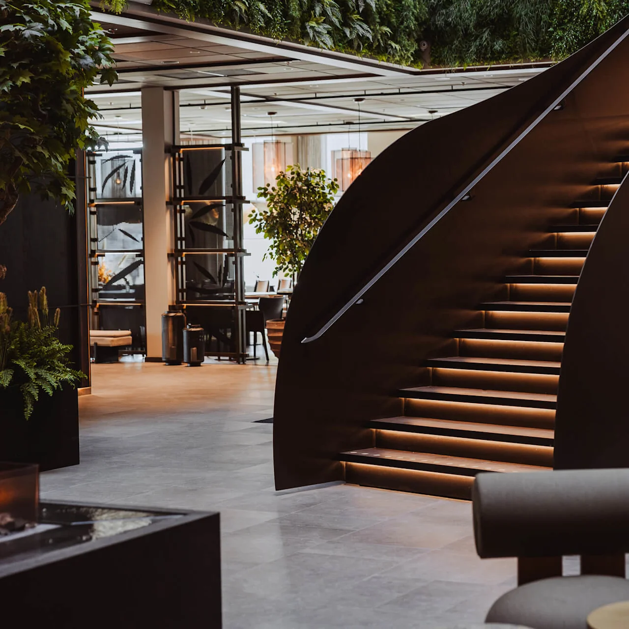 En modern trappa med stämningsbelysning stiger elegant i en stilfull, växtfylld interiör med sittplatser och dekorativa element.