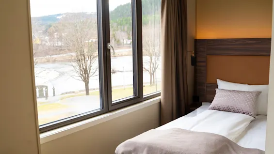 A cozy hotel room with a comfortable bed and a large window offering a scenic view of a river and trees.