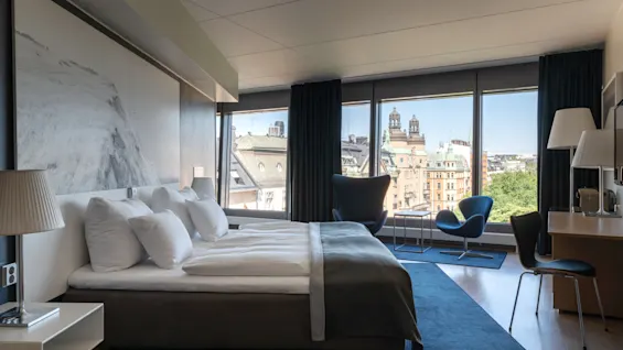 A modern hotel room featuring a large bed, a desk area, and a seating area by a window with a city view.