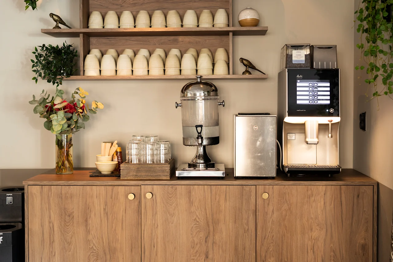 En självbetjäningsstation med kaffemaskin som erbjuder dryckesval, en vattendispenser och koppar. PAPPERSKORG.
