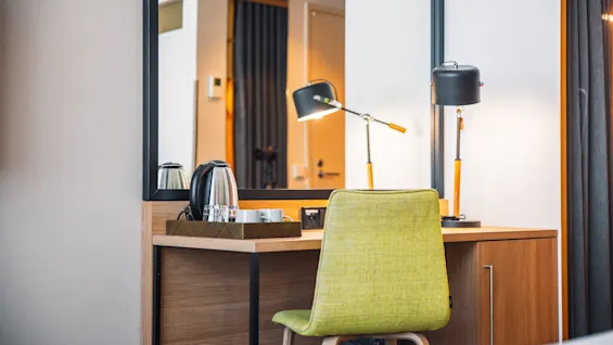 A wooden desk with a lime green chair and two black lamps arranged beside a kettle and cups. The setup is in a modern room with a mirror and curtain backdrop.