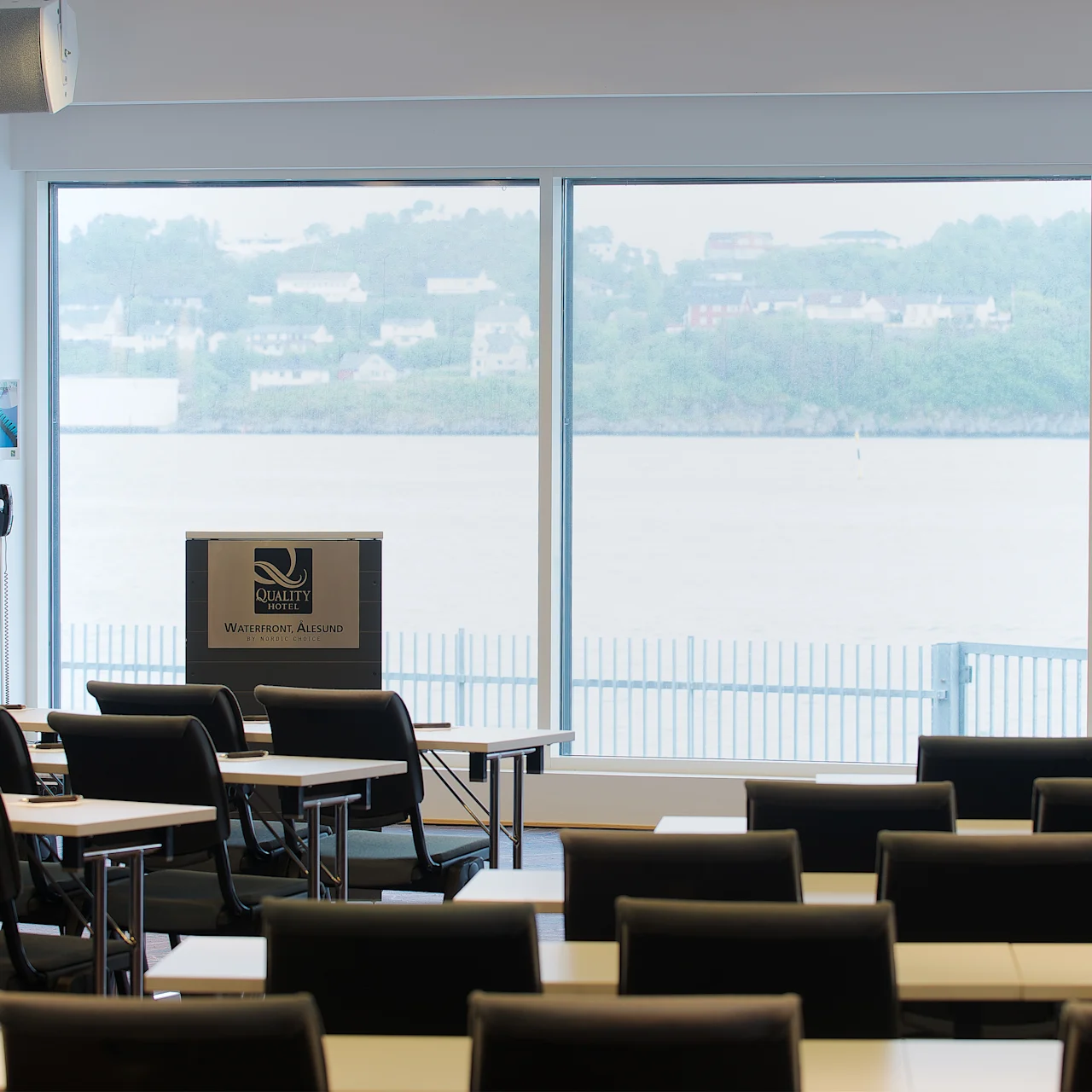 Ett podium märkt "Quality Hotel Waterfront Ålesund by Nordic Choice" står i en konferenssal med rader av tomma stolar, vänd mot stora fönster med utsikt över vattnet och en avlägsen sluttning.