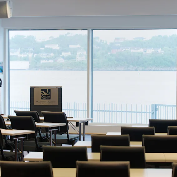 En talerstol merket "Quality Hotel Waterfront Ålesund by Nordic Choice" står i en konferansesal med rader av tomme stoler, vendt mot store vinduer med utsikt over vannet og en fjern åsside.
