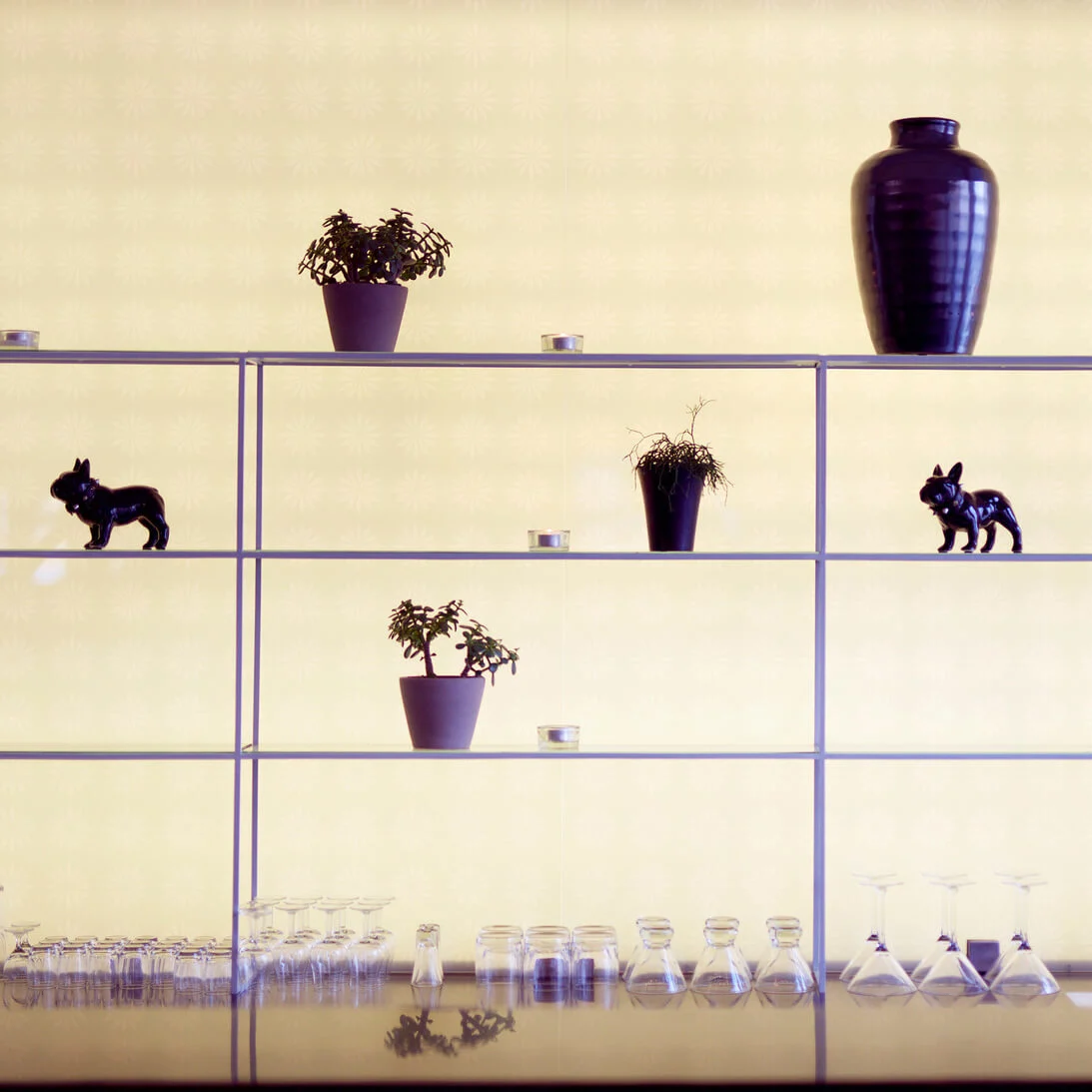 A shelving unit holds assorted items like plants, decorative vases, and candle holders against a patterned wallpaper, next to a window with sheer curtains.