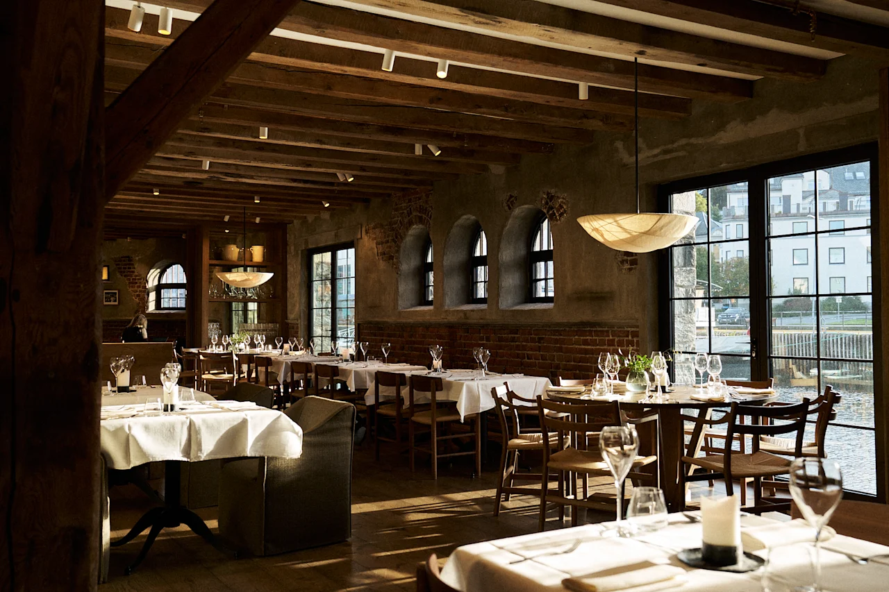 Et varmt og indbydende restaurantinteriør på Sjøbua, Hotel Brosundet, med dækkede borde, træbjælker og store vinduer med udsigt over vandet.