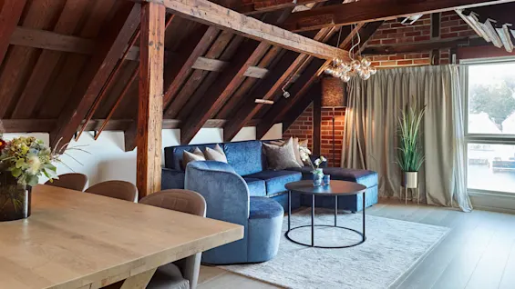 A cozy attic living room with a blue sofa, exposed wooden beams, and a brick wall. A modern chandelier illuminates the space.