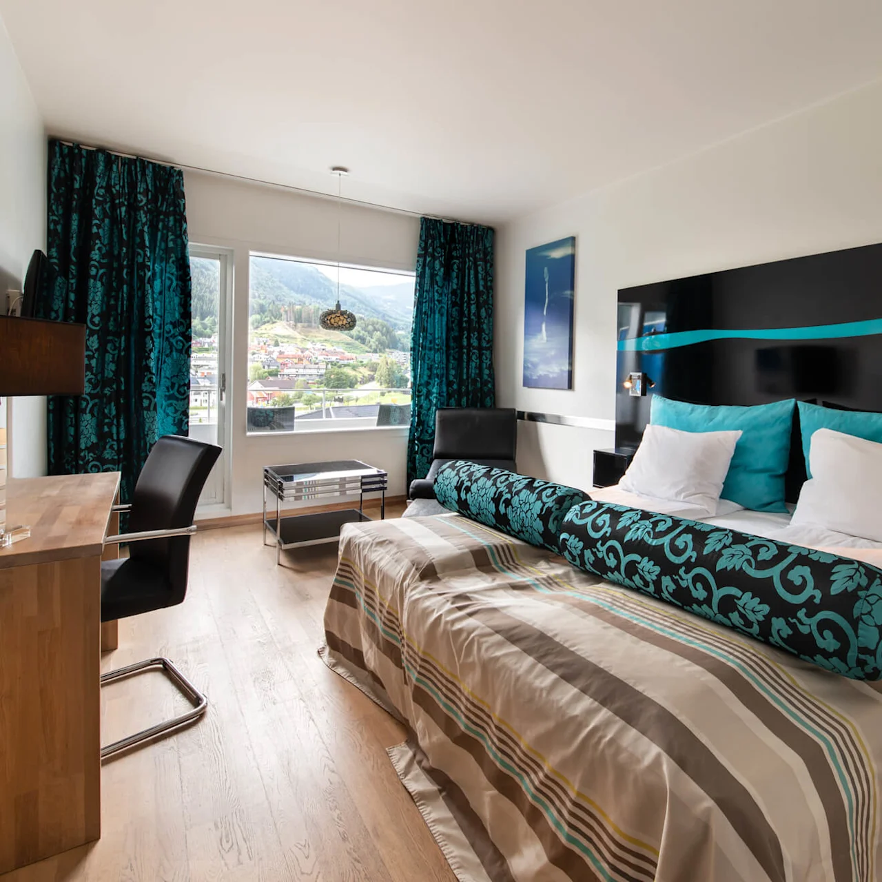 En snyggt bäddad säng med turkosa och vita kuddar står i ett modernt hotellrum. Ett skrivbord, stol och fönster med bergsutsikt kompletterar den stilrena inredningen.