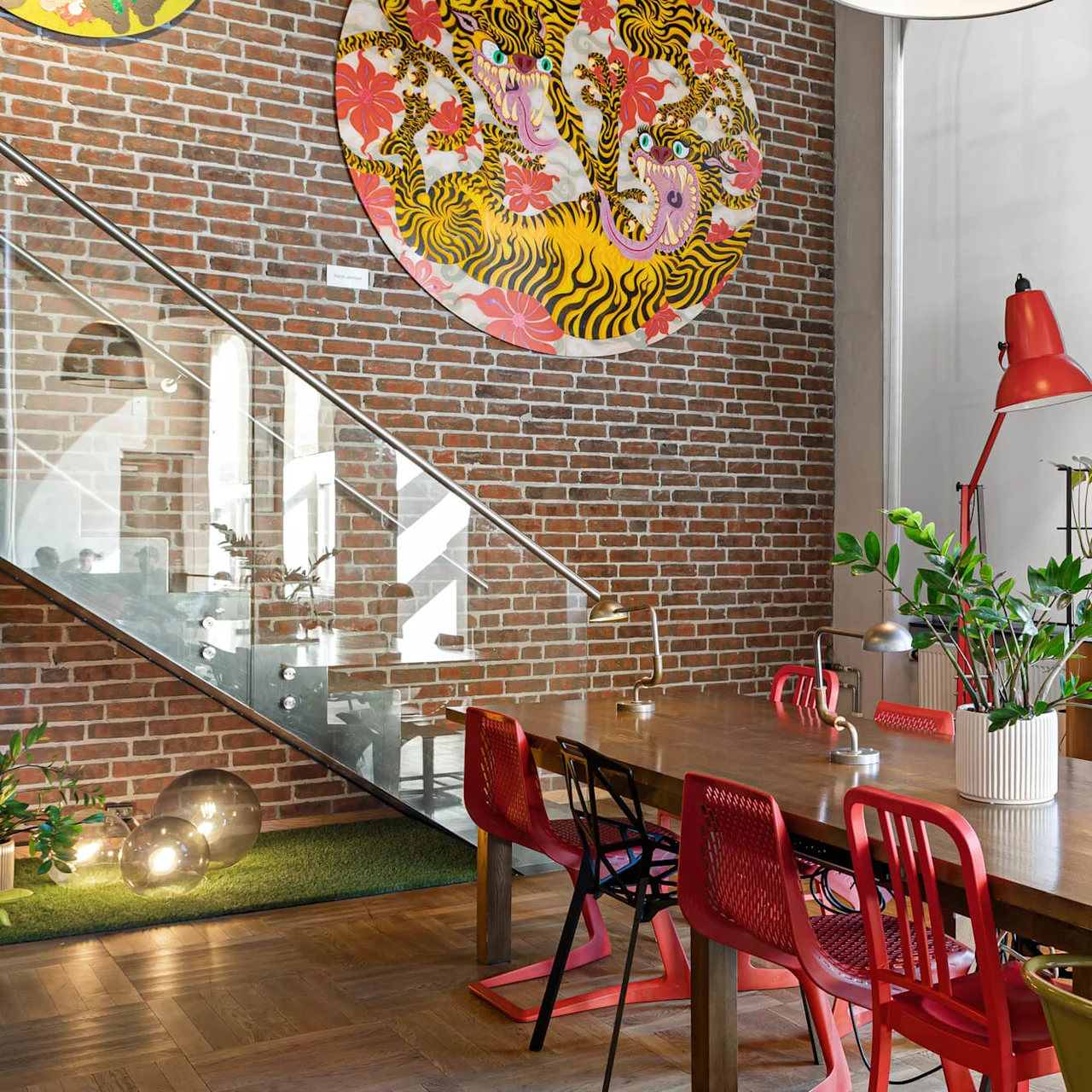 A wooden table surrounded by colorful chairs is adorned with a plant and desk lamps. In a room with exposed brick walls, vibrant wall art, and a glass staircase.
