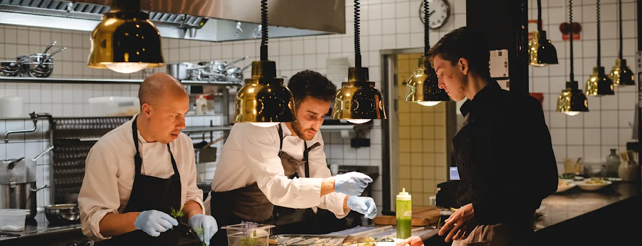 Kokker som forbereder retter på en benk, iført forklær og hansker, i et profesjonelt kjøkken med hengende lamper og flislagte vegger. Tallerkener arrangert på rad, hver med mat.