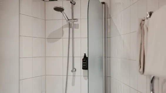 Et lyst, flisebelagt brusebad med en glasskærm, brusehoved og en flaske BODY WASH. Friske håndklæder hænger på en hylde.