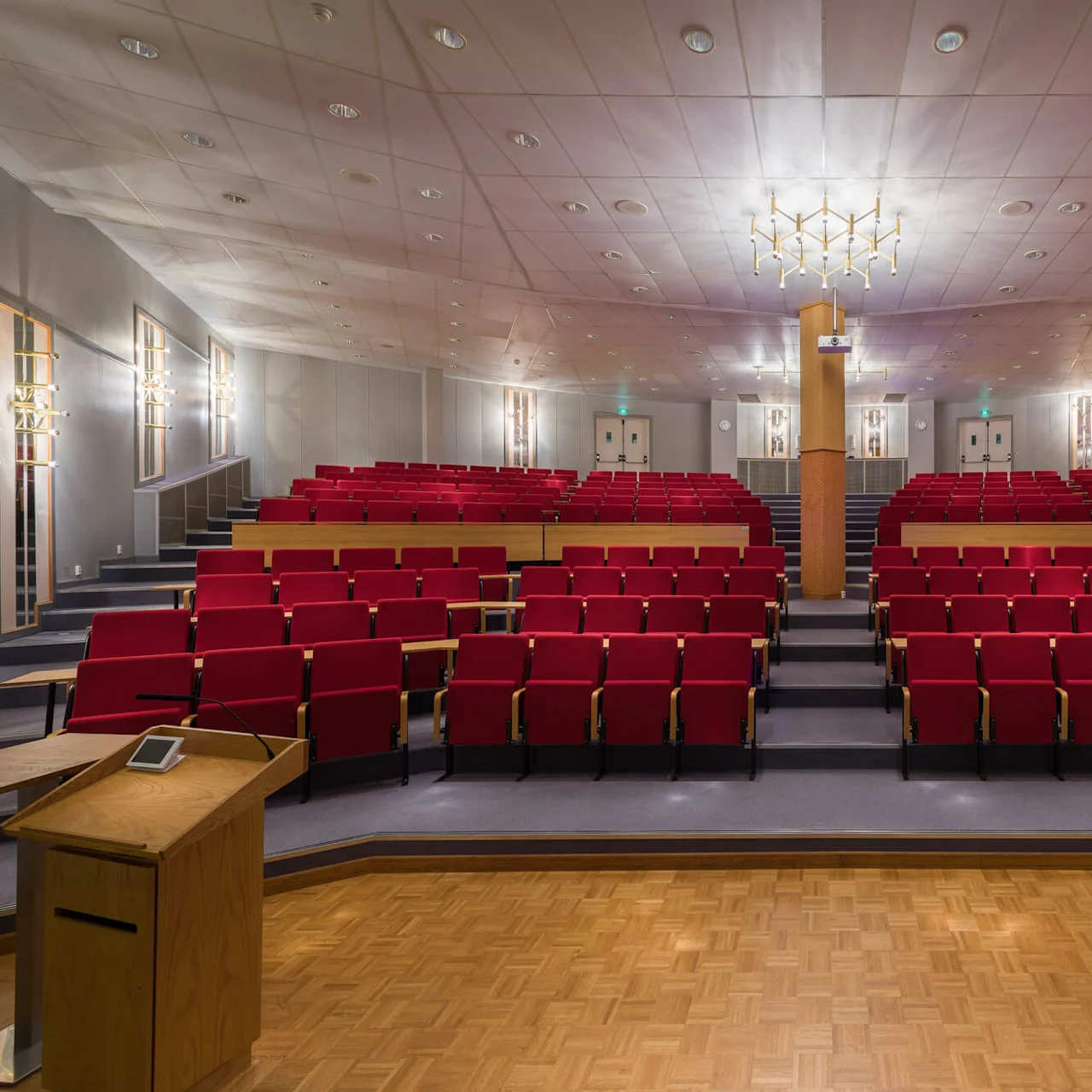 Et træpodium står på en scene med en mikrofon, mod røde auditoriepladser i en lys foredragshal med hvidt loft.