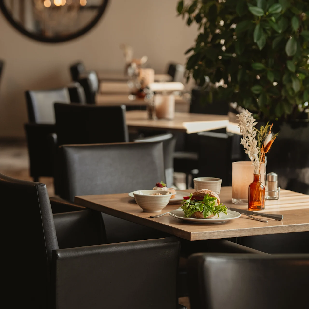 A dining table holds a salad and bowl beside a glass and vase in a restaurant with black chairs, soft lighting, and a large plant nearby.