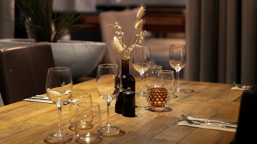 Ett träbord är dukat med tomma vinglas, bestick på servetter och en vas med torkade blommor, vilket skapar en elegant middagsscen med dämpad belysning.