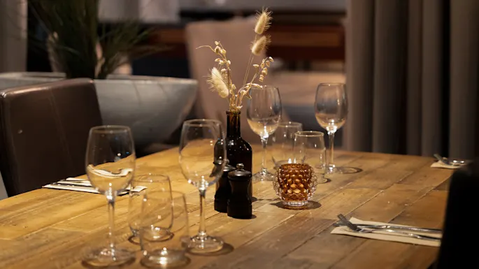 Et trebord er dekket med tomme vinglass, bestikk på servietter og en vase med tørkede blomster, som skaper en elegant middagsstemning med dempet belysning.