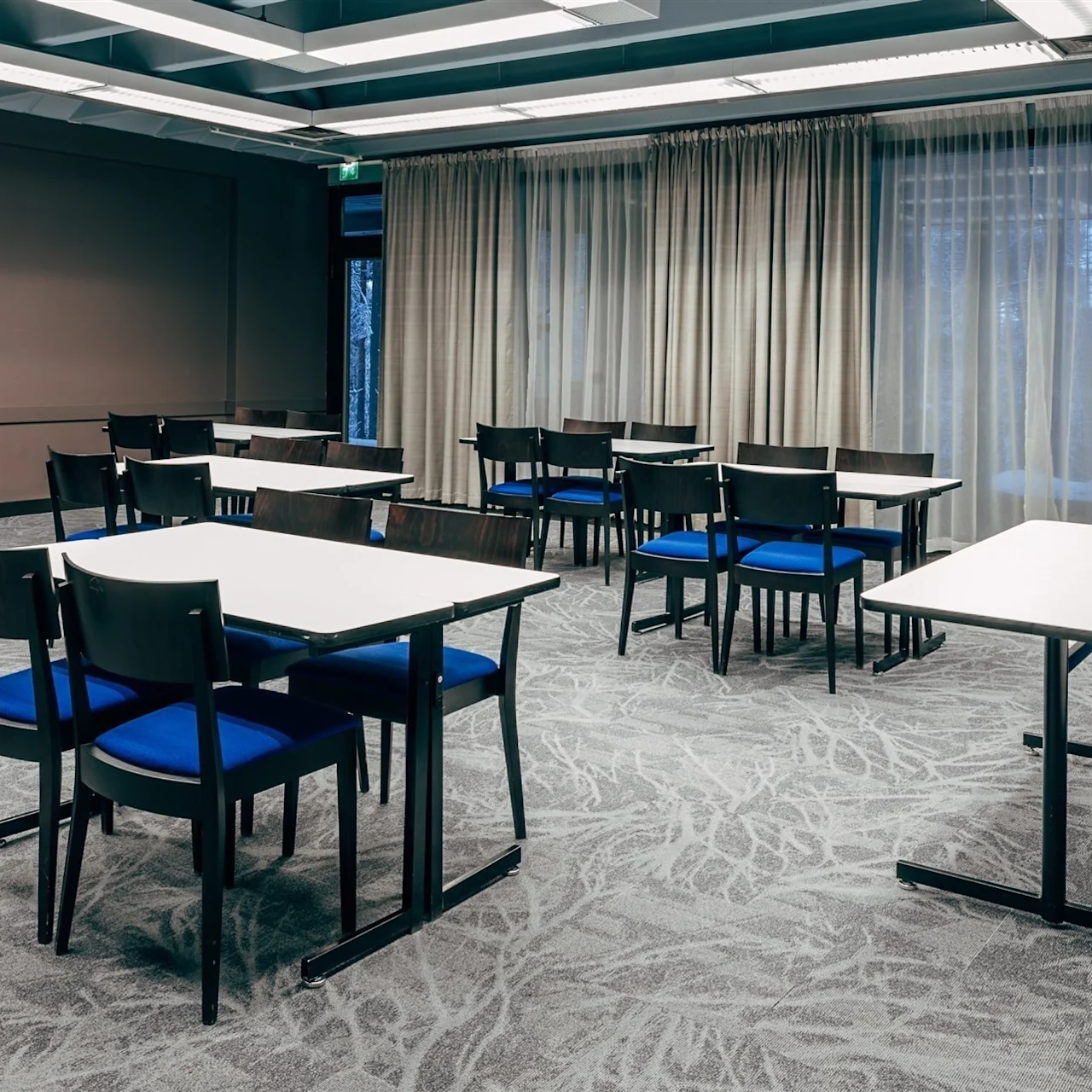 Tables with chairs are arranged casually in a carpeted room. A clock is on the wall and curtains cover the windows. A flip chart stands near a window.