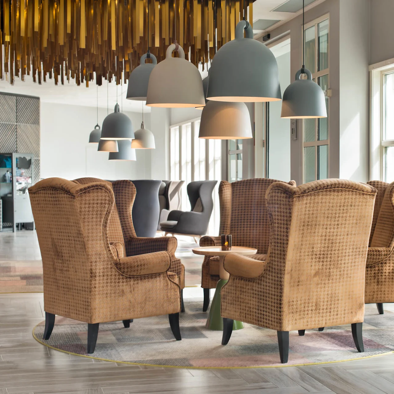 Brown upholstered armchairs encircle a small wooden table beneath pendant lights, in a bright lounge with large windows and a decorative ceiling.