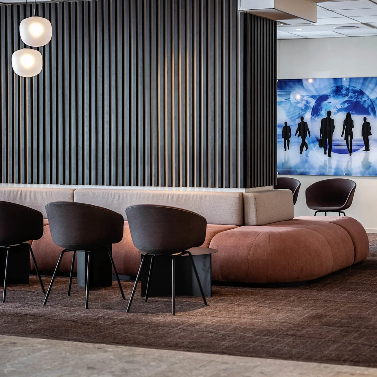 Et siddeområde har moderne stole og en sofa, der vender mod en træpanel. Et digitalt kunstværk med silhuetter og en globus pryder væggen ved siden af en tændt pejs.