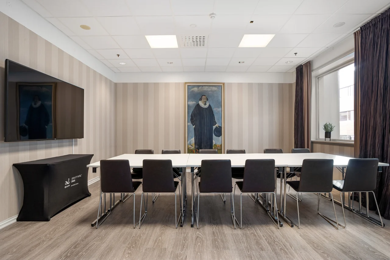Et stort bord omgivet af sorte stole i et mødelokale. Et portræt hænger på væggen. Et fladskærms-TV er monteret til venstre. Beige-stribede vægge tilføjer et formelt touch.