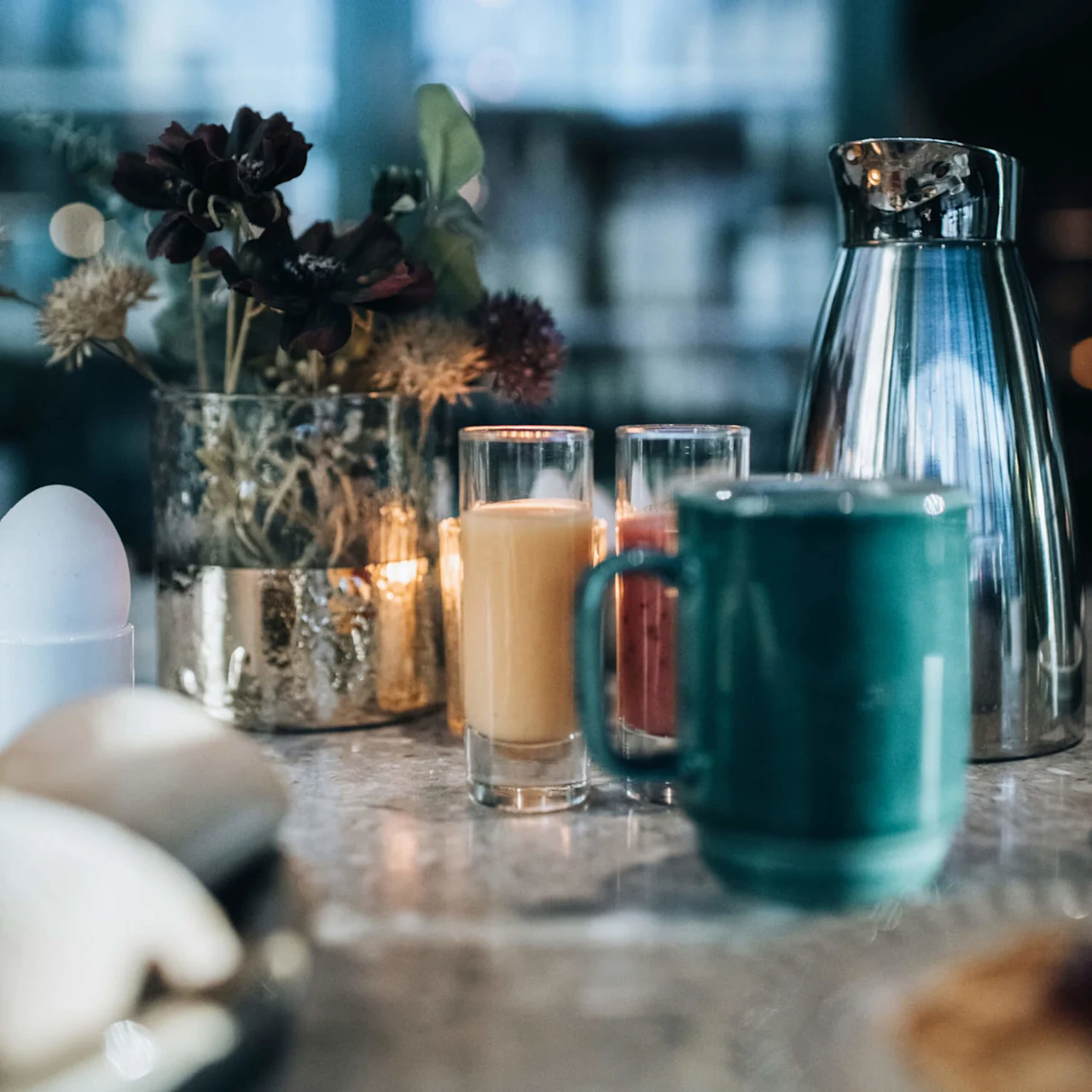 En turkos mugg står på en marmorbänk, omgiven av en metalltermos, en blomvas, ljuslyktor och ett äggbägare, vilket skapar en mysig frukoststämning.