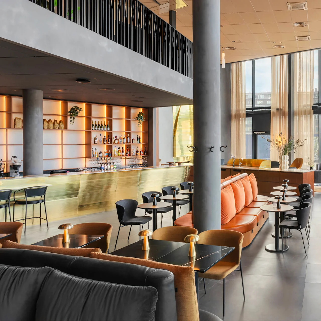 Ett stilfullt, modernt loungeområde med orange soffor och svarta bord i en rymlig miljö med stora fönster och grönska. En välfylld bar med belysta hyllor sprider värme.