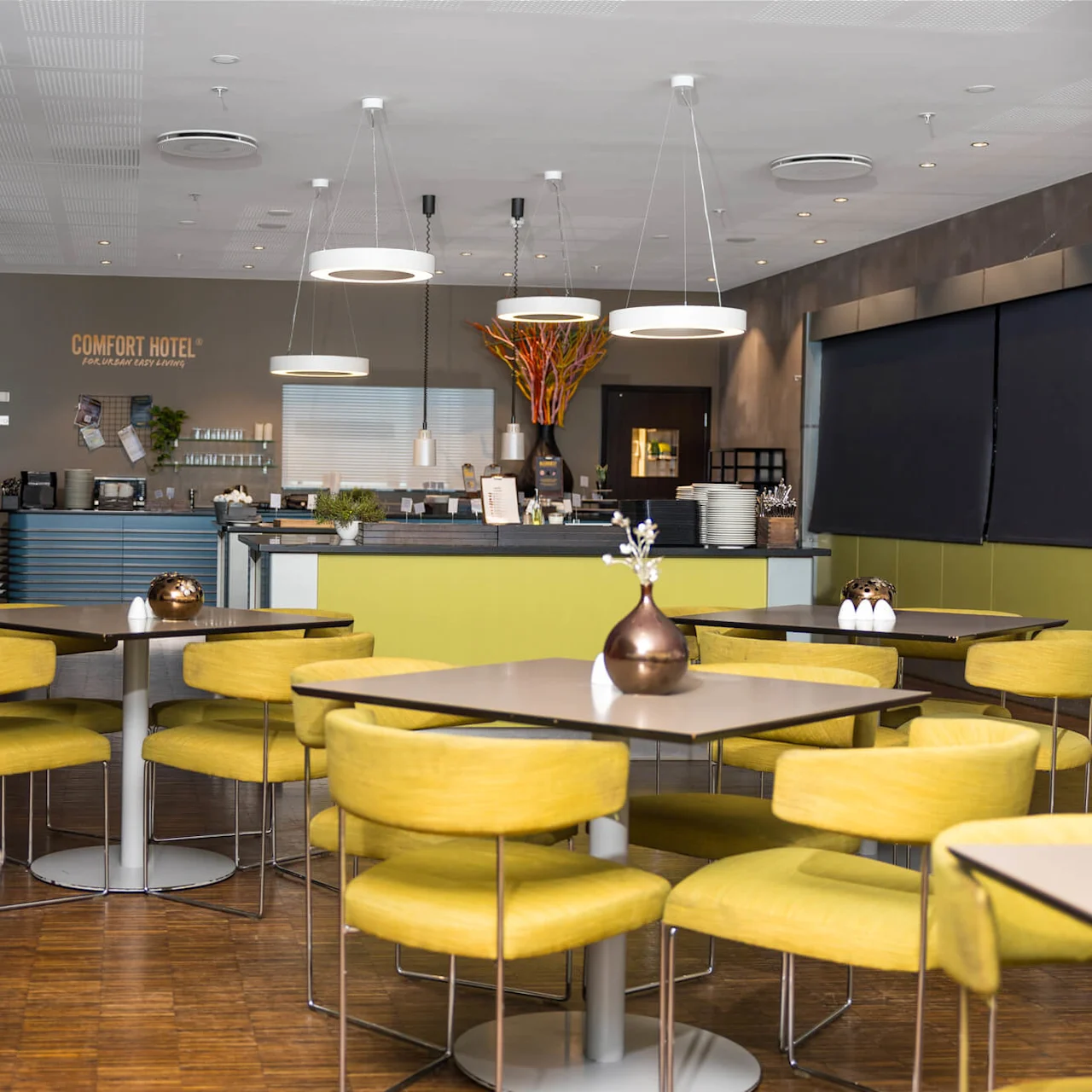 Tables with yellow chairs are set in a modern, well-lit dining area. Large plants and abstract decorations fill the space. Text: "COMFORT HOTEL™ For Dream Easy Living."