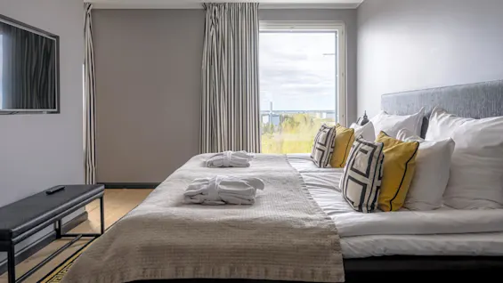 A neatly made bed with patterned pillows and two folded robes placed on it, positioned beside a window with striped curtains and overlooking a leafy view. A TV mounted on the wall.