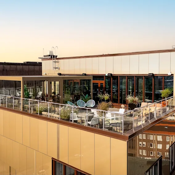 Hotelltaksterrass med uteservering, växter och utsikt över staden. Hotellskylt synlig.