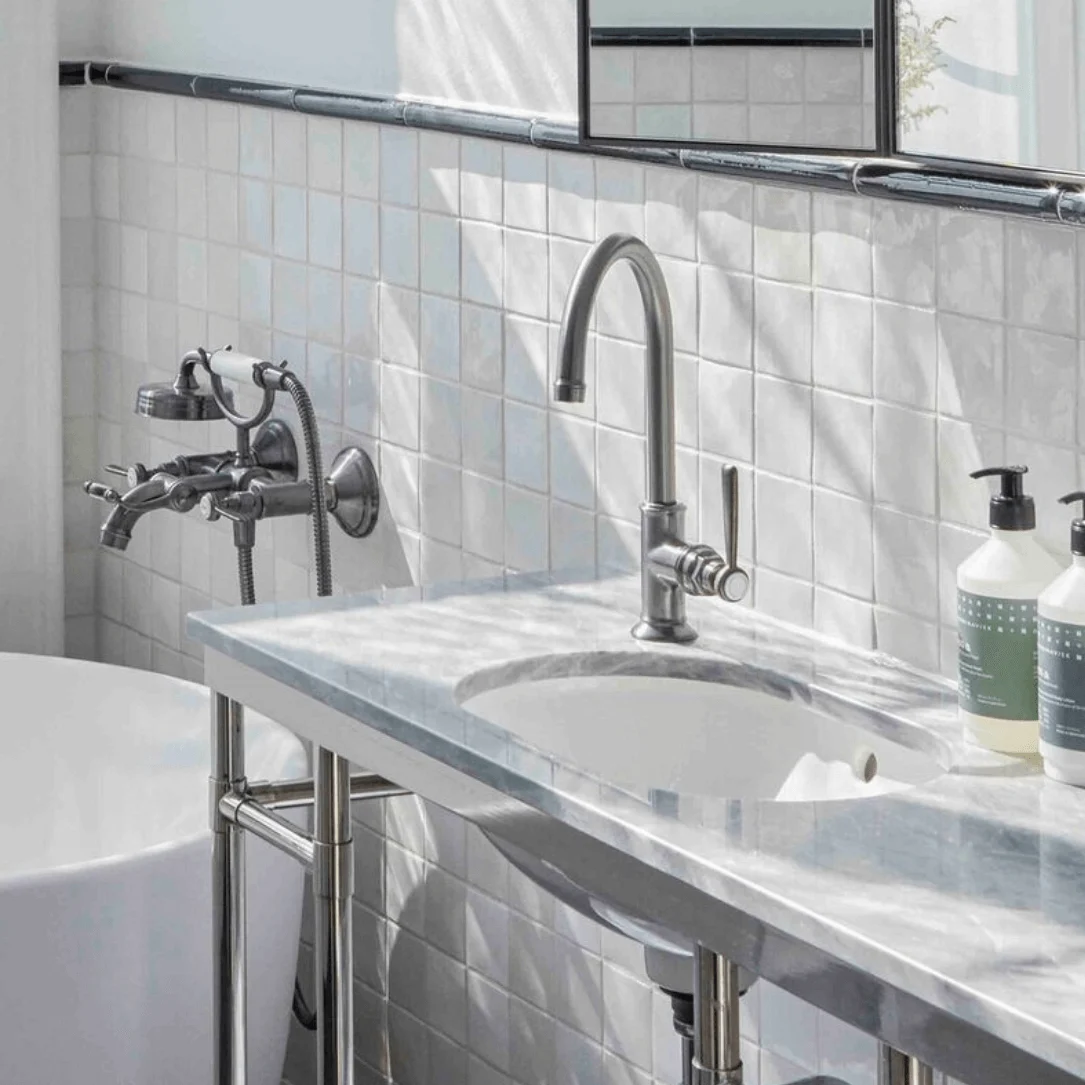 A white bathtub and sink with a chrome faucet are set against tiled walls, with toiletries on the windowsill and countertop. Light streams through a partially open window. 