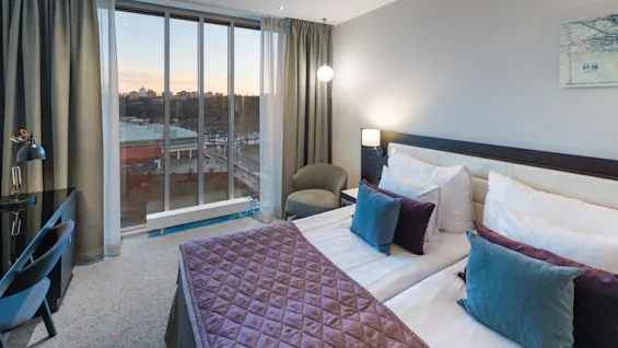 En seng med lilla og teal puter står i et moderne hotellrom, vendt mot et stort vindu med utsikt over et bybilde ved solnedgang.