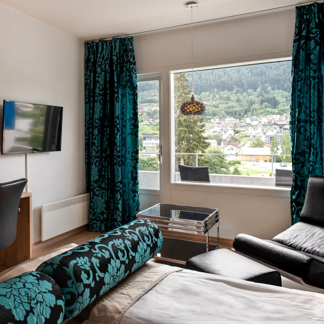 Ett modernt hotellrum med ett skrivbord med en lampa, en väggmonterad TV och ett stort fönster med utsikt över en stad och skogsklädda kullar. Blå mönstrade gardiner lägger till färg.
