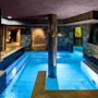 En indendørs pool med klart blåt vand omgiver en central søjle, omgivet af stenvægge og omgivende belysning. Døre og vinduer med udsigt til naturen forbedrer den rolige atmosfære.
