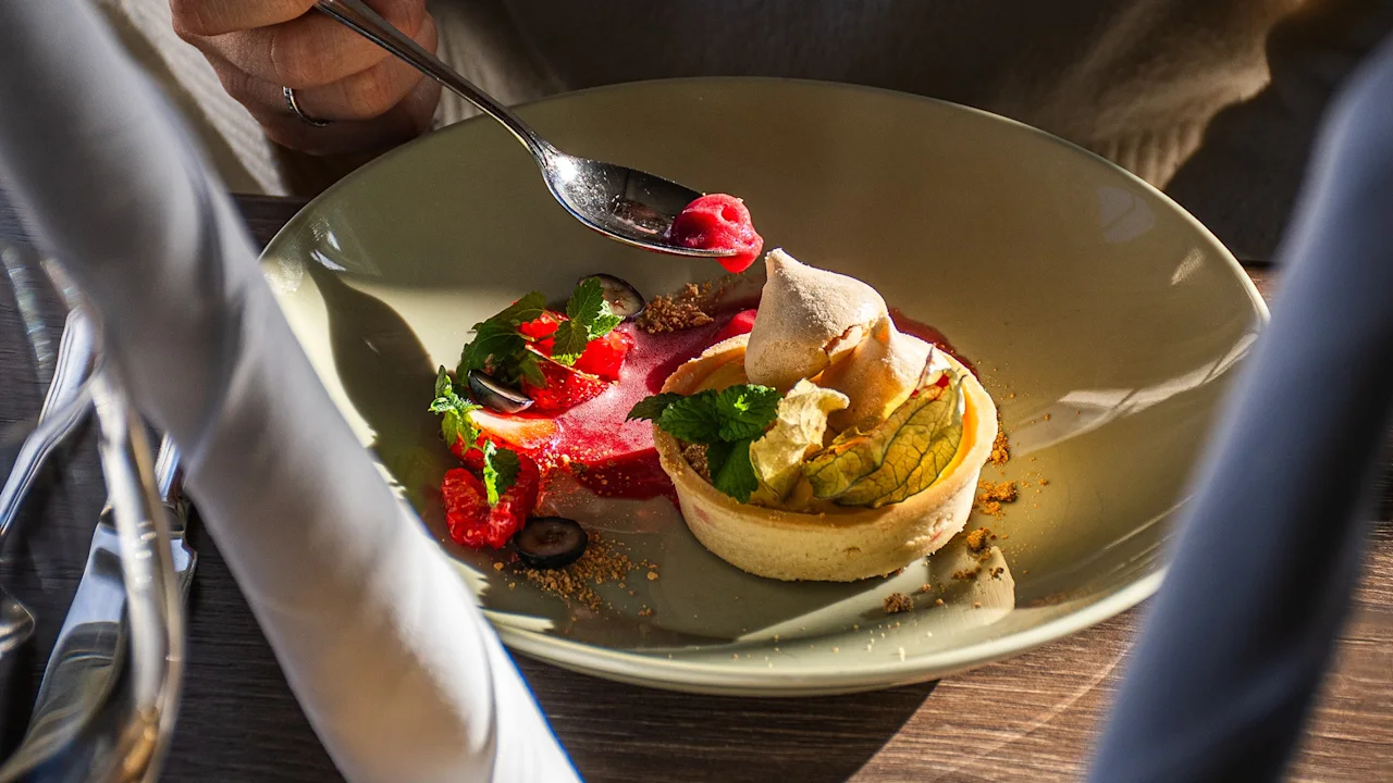 En person njuter av en vackert upplagd efterrätt med en sked, med en tårta, bär och en röd sås.