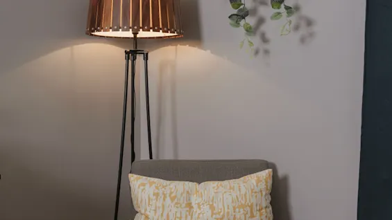 A gray chair with a yellow patterned pillow sits under a tall floor lamp. Green plants hang on a light gray wall in a cozy corner with wooden flooring.