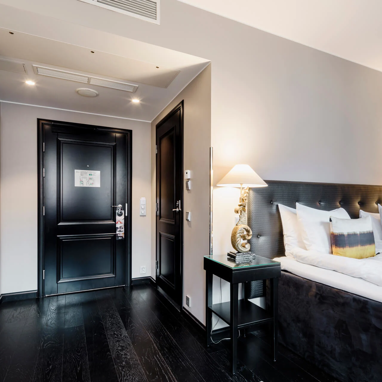 En pent sengeredning med hvite puter og mørk sengegavl står ved siden av to stilige lamper på et elegant hotellrom med mørkt tregulv og lyse vegger.