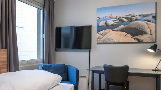 Et velholdt moderne værelse indeholder en seng, en blå stol, et skrivebord med lampe og et vægmonteret TV. Et stort vindue og et kystlandskab maleri giver sammenhæng.