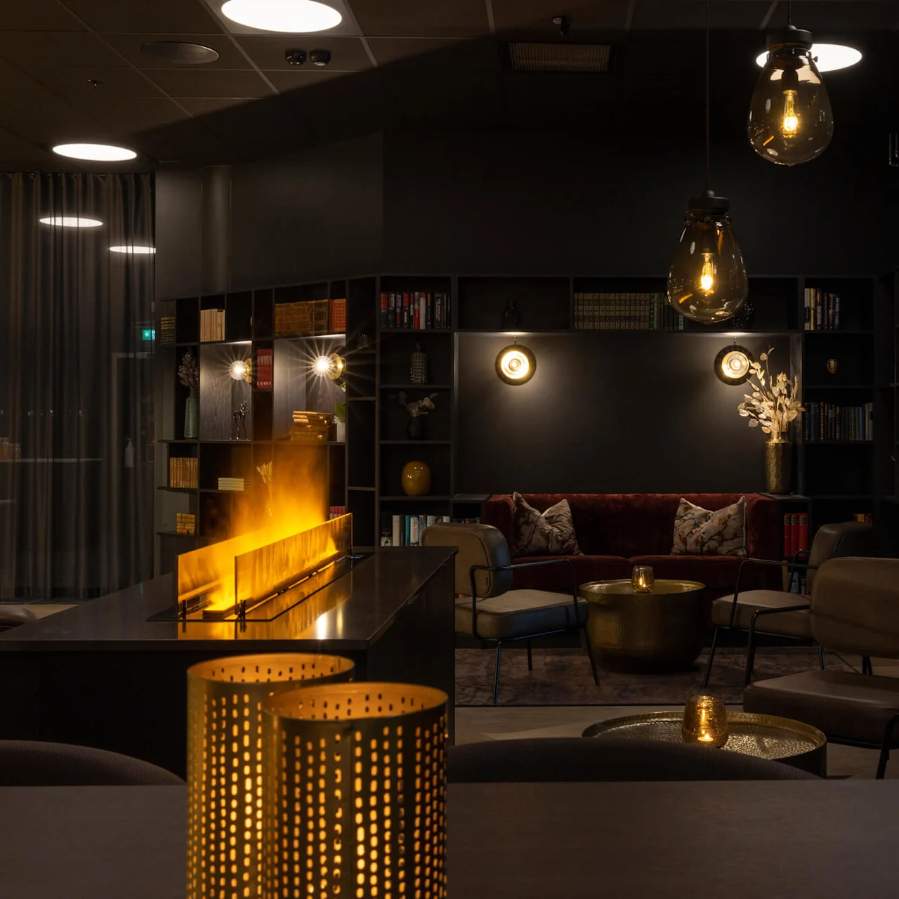 Cozy modern lounge with dim lighting, a lit fireplace, plush seating, bookshelves, and decorative lamps.
