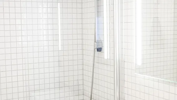 Badkar med duschhuvud monterat på en kaklad vägg, omgivet av tre flaskor. Vikta handdukar ligger på en svart bänk bredvid ett handfat i ett ljust, rent badrum.