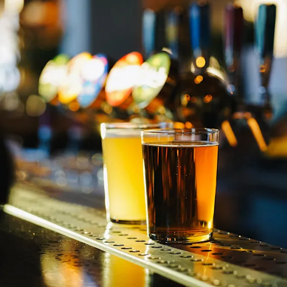 Two glasses of beer sit on a bar counter; one light, the other dark. Bar taps with colorful handles are visible in the blurred background.