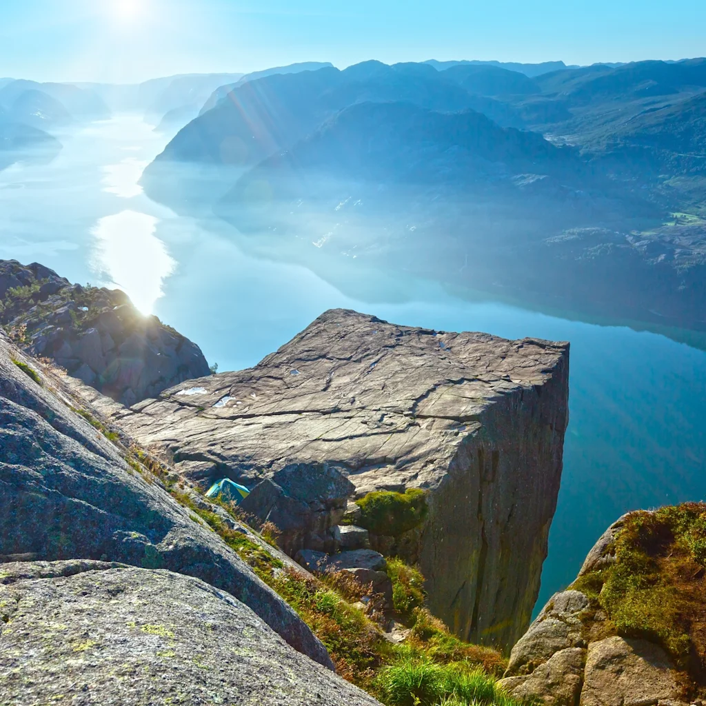 En flat fjellformasjon stikker ut over en blå fjord, omgitt av robuste fjell under en klar himmel, med sollys som reflekteres i vannet nedenfor.