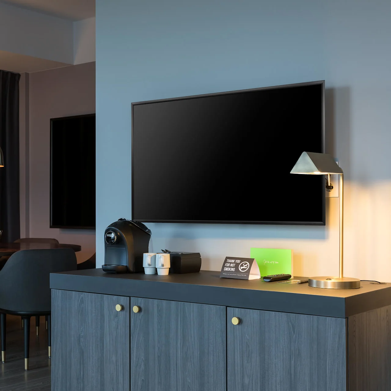 TV på en vegg, med kaffemaskin og lampe på et skap i nærheten. Bak, et bord med stoler under hengende belysning. Et skilt på skapet sier, "THANK YOU FOR NOT SMOKING".