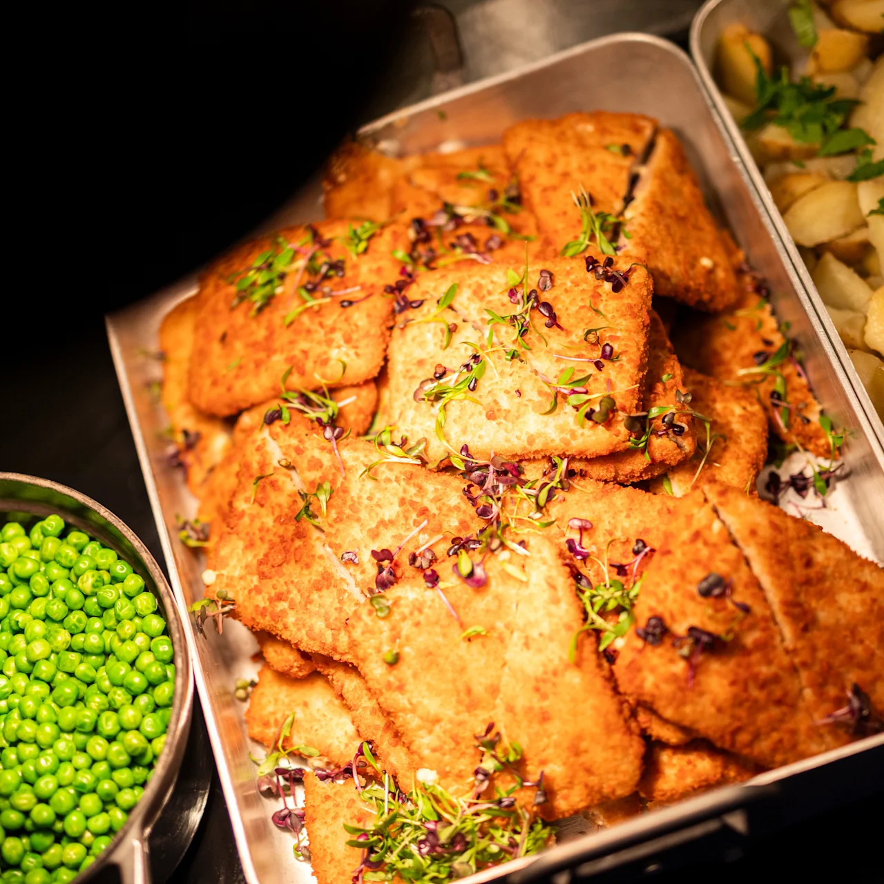 Sprøde panerede fiskefileter pyntet med urter i en metalform, ledsaget af en skål med grønne ærter til venstre og stegte kartofler med persille til højre.