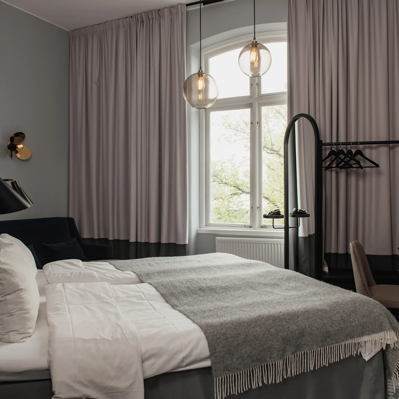 En prydligt bäddad säng med grått sängkläder står i ett modernt, stilrent sovrum. Gardiner täcker ett stort fönster; ett skrivbord, en stol och en klädhängare kompletterar den mysiga, minimalistiska inredningen.