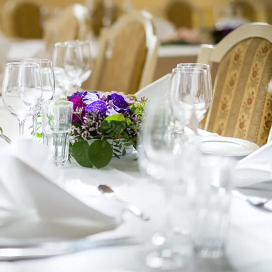 Elegant spisebord dækket med foldede hvide servietter og glas i en formel setting. Lilla blomster i midten tilføjer farve, omgivet af udsmykkede stole i en raffineret banketsal.
